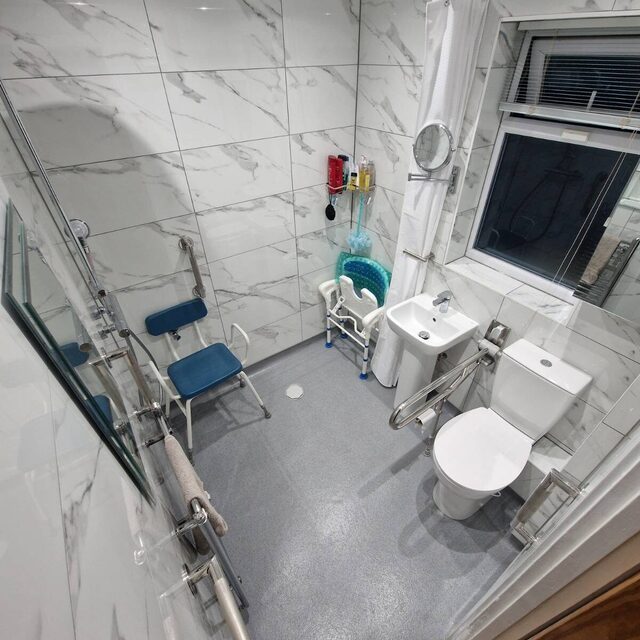 Accessible wet room fitted with stainless steel grab rails, a fold-down shower seat, comfort-height toilet and non-slip safety flooring, installed by Downs Plumbing & Heating