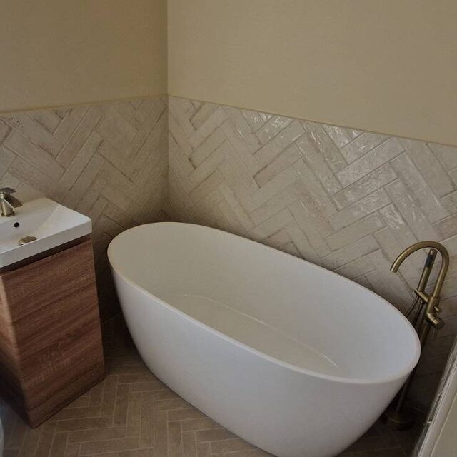 Freestanding oval bath fitted alongside a close-coupled toilet and walnut vanity unit, with herringbone tiled walls and a brushed gold bath filler, installed by Downs Plumbing & Heating