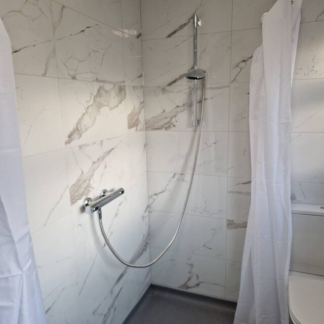 Level-access wet room shower area with marble-effect tiling, a thermostatic bar mixer on a riser rail and a floor drain, installed by Downs Plumbing & Heating