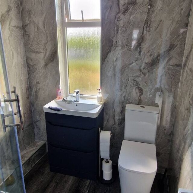 Bathroom finished in marble-effect wall panelling with a navy blue vanity unit, close-coupled toilet and dark wood-effect flooring, fitted by Downs Plumbing & Heating