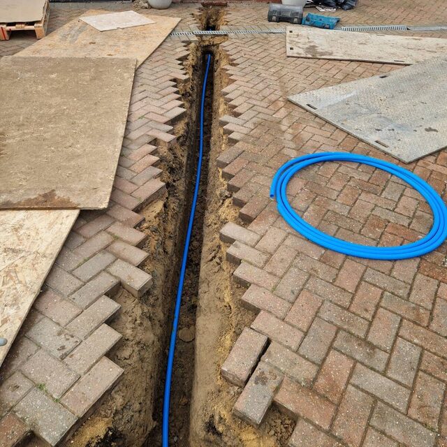 New blue MDPE water main laid in a trench across a block-paved driveway, replacing an old supply pipe, carried out by Downs Plumbing & Heating