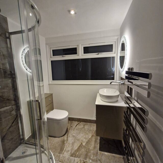 Bathroom with a curved quadrant shower enclosure, round LED mirror, wall-hung basin and marble-effect floor tiles, fitted by Downs Plumbing & Heating